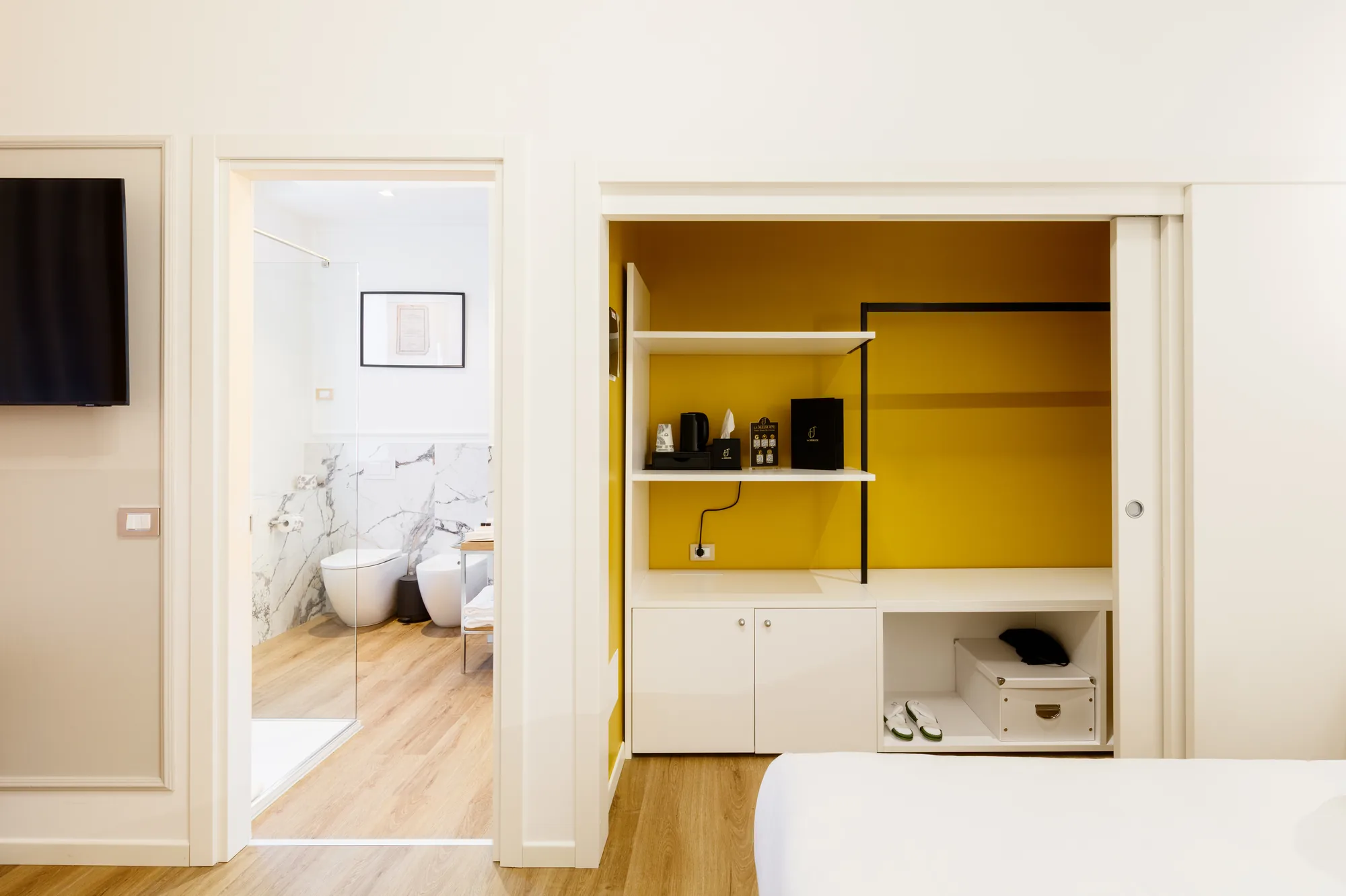 The image shows a wardrobe with white shelves and a yellow wall, containing a kettle and toiletries, with a door leading to a modern bathroom