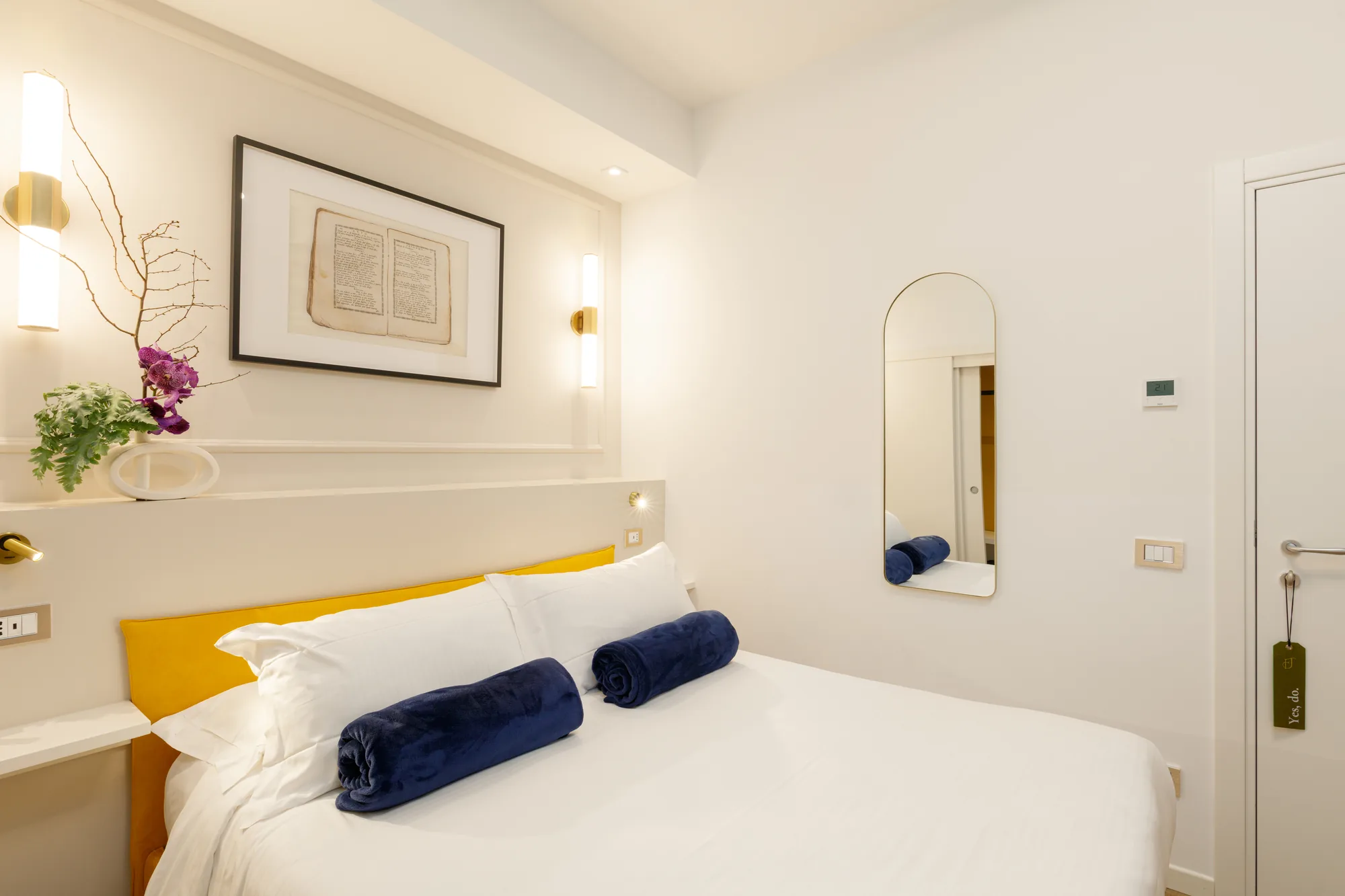 Modern bedroom with a double bed, blue blankets, and minimalist decorations, including a painting and a vase of flowers