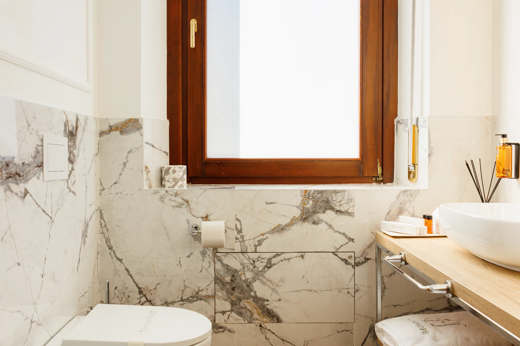 Alt text: Interno di un bagno elegante con pareti in marmo, una finestra in legno e accessori per la cura personale
