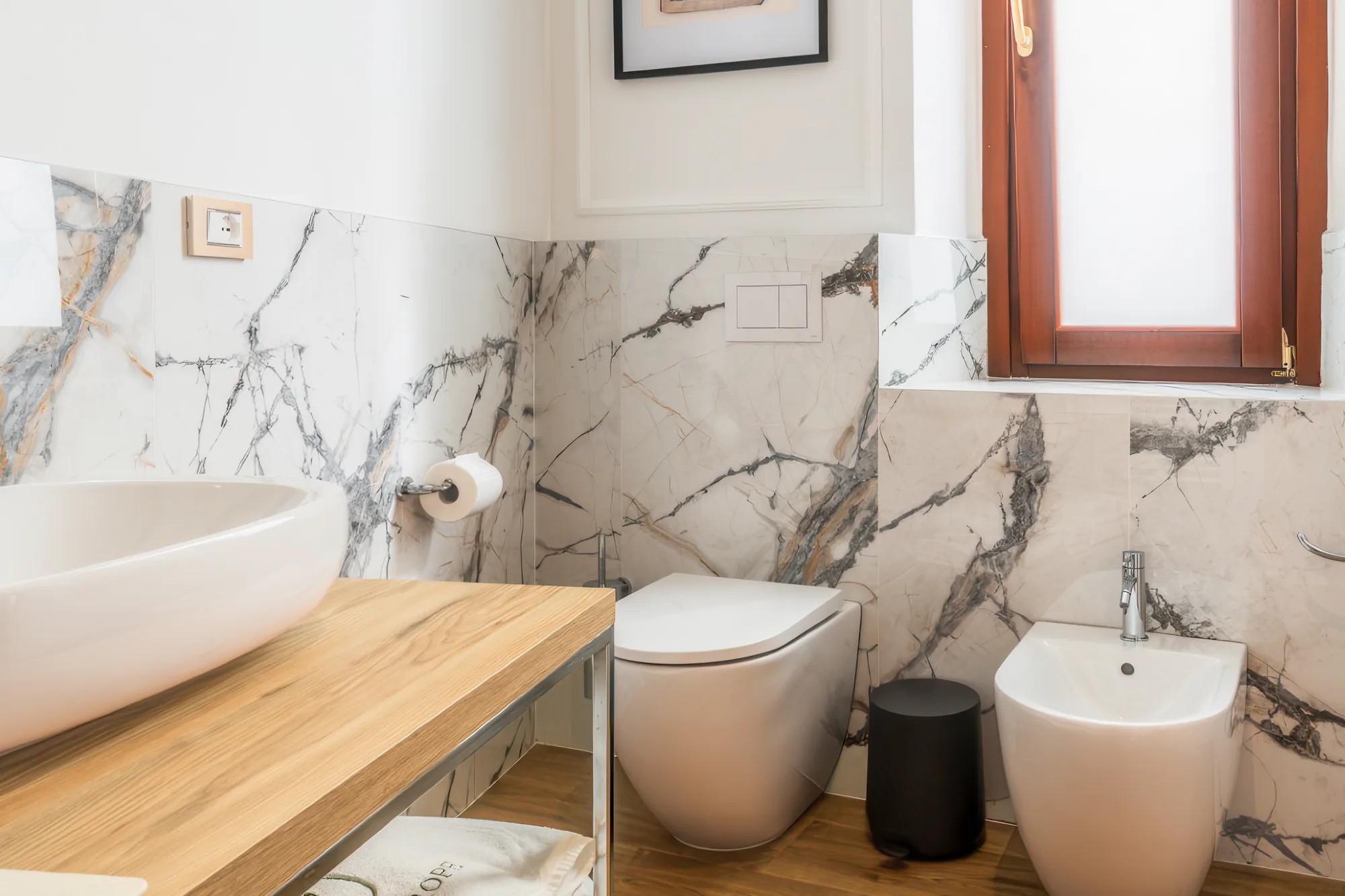 Bagno moderno con pareti in marmo, lavabo bianco su un piano in legno, wc e bidet, e una finestra con infissi in legno