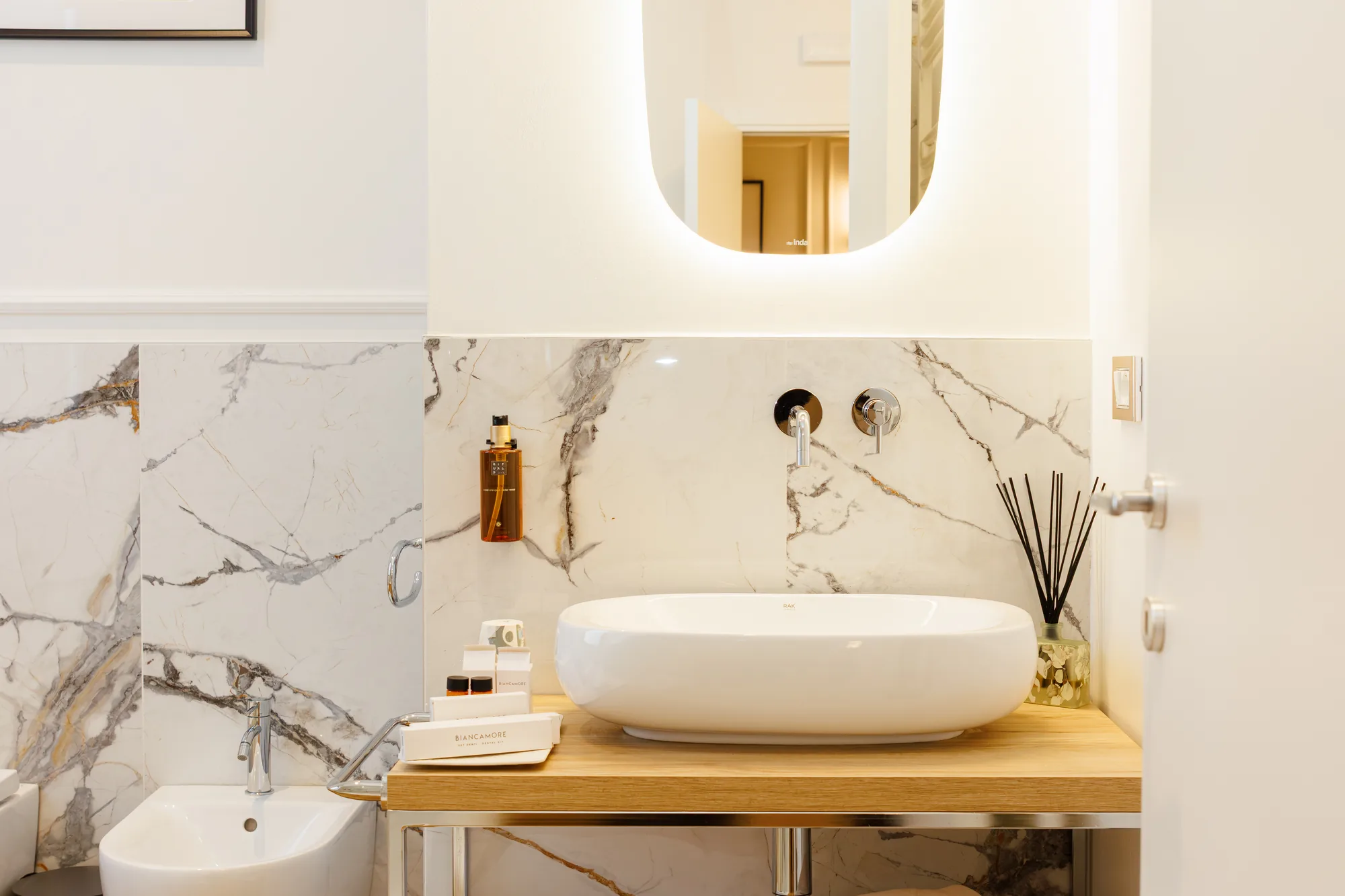 A modern bathroom with marble walls, white sink on a wooden countertop, personal care products, and an aroma diffuser.