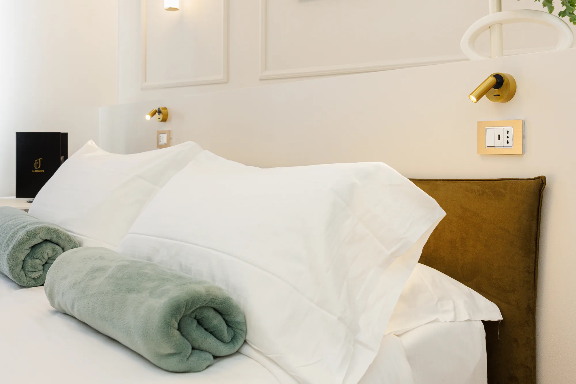 Immagine di un letto con coperte arrotolate e cuscini bianchi, illuminato da una lampada da parete e con una presa elettrica visibile