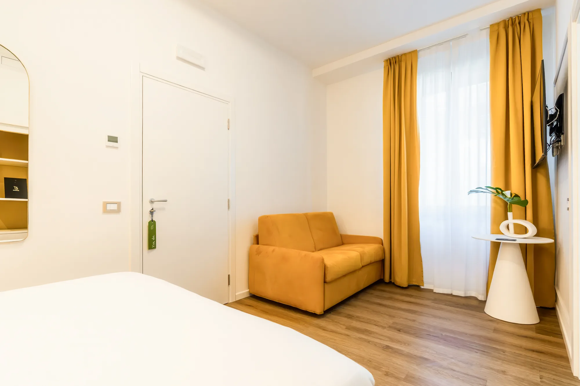 A hotel room with a yellow sofa, a window with yellow curtains, a white coffee table, and a door.