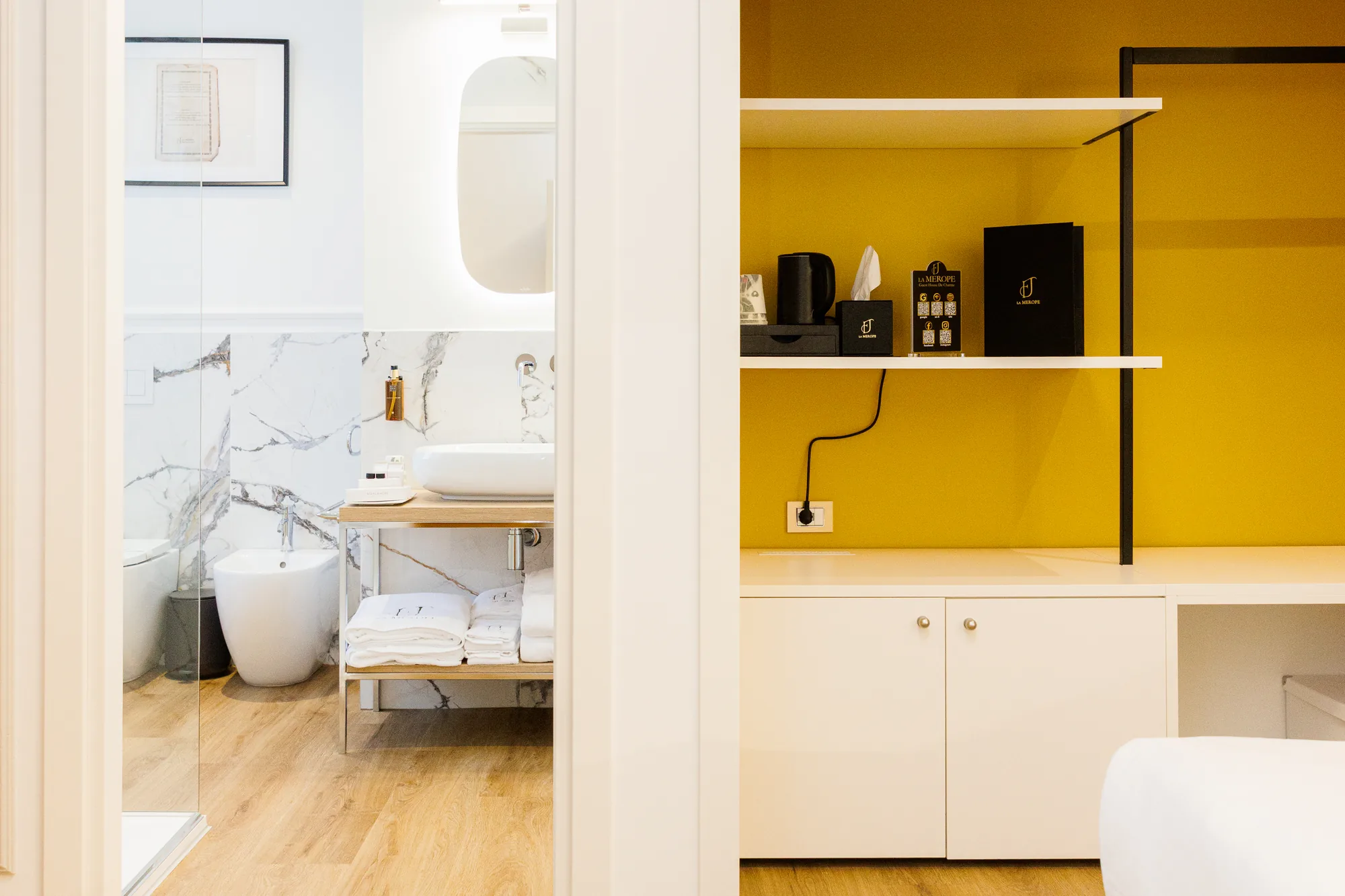 View of a modern bathroom with marble walls, sink on a wooden cabinet, and folded towels, next to an area with a coffee machine and products.
